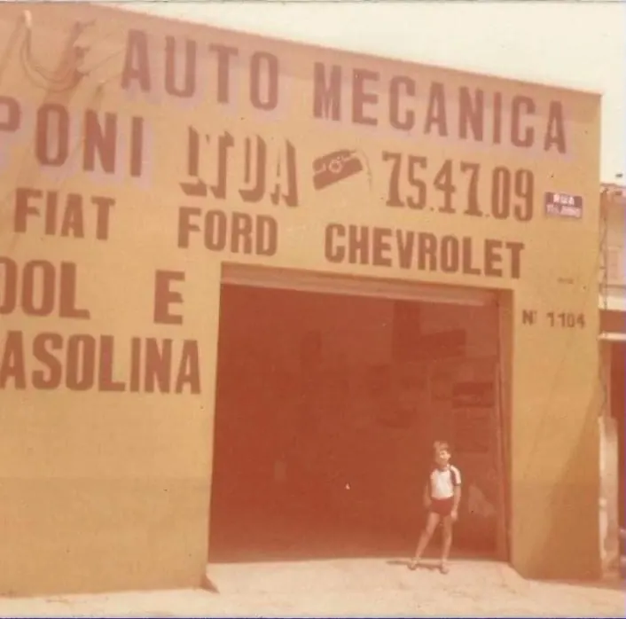 Fotografia antiga e desbotada: fachada ocre com letreiro pintado AUTO MECANICA PONI LTDA, telefone 7547.09, as linhas FIAT FORD CHEVROLET e ÁLCOOL E GASOLINA, e um menino de camiseta branca na porta