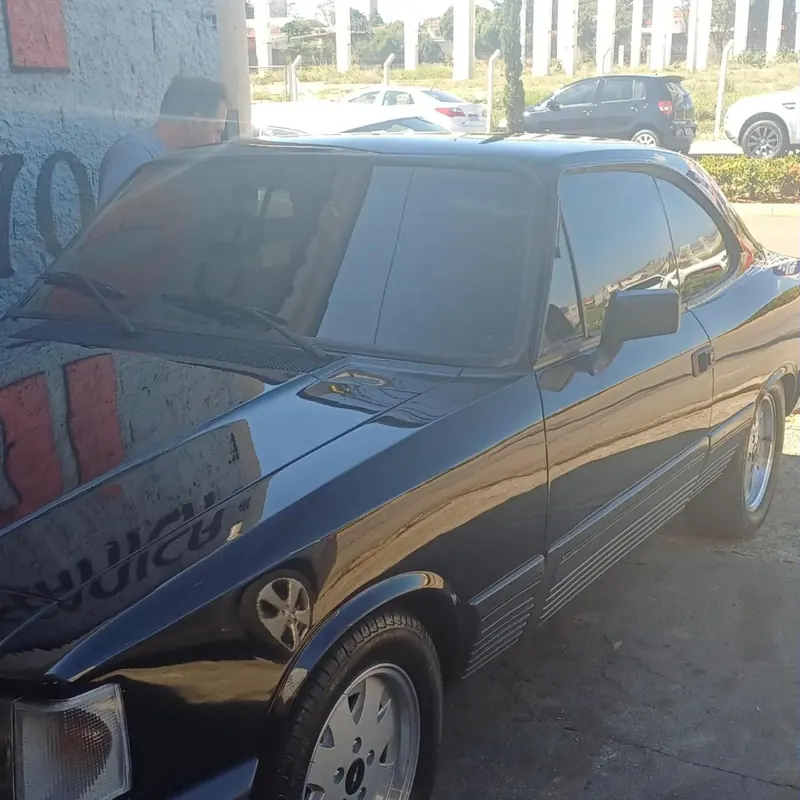 Chevrolet Opala preto estacionado na calçada da oficina, com o letreiro da parede refletido na lataria